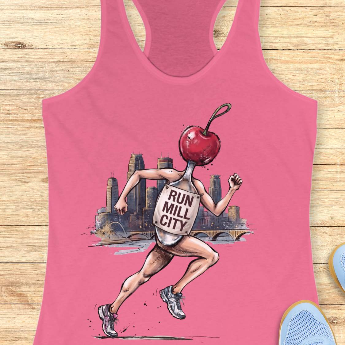 A pink racerback tank top laid flat on a light wood background, featuring a graphic of a running spoon with a cherry for a head and the words 'RUN MILL CITY' on its body.