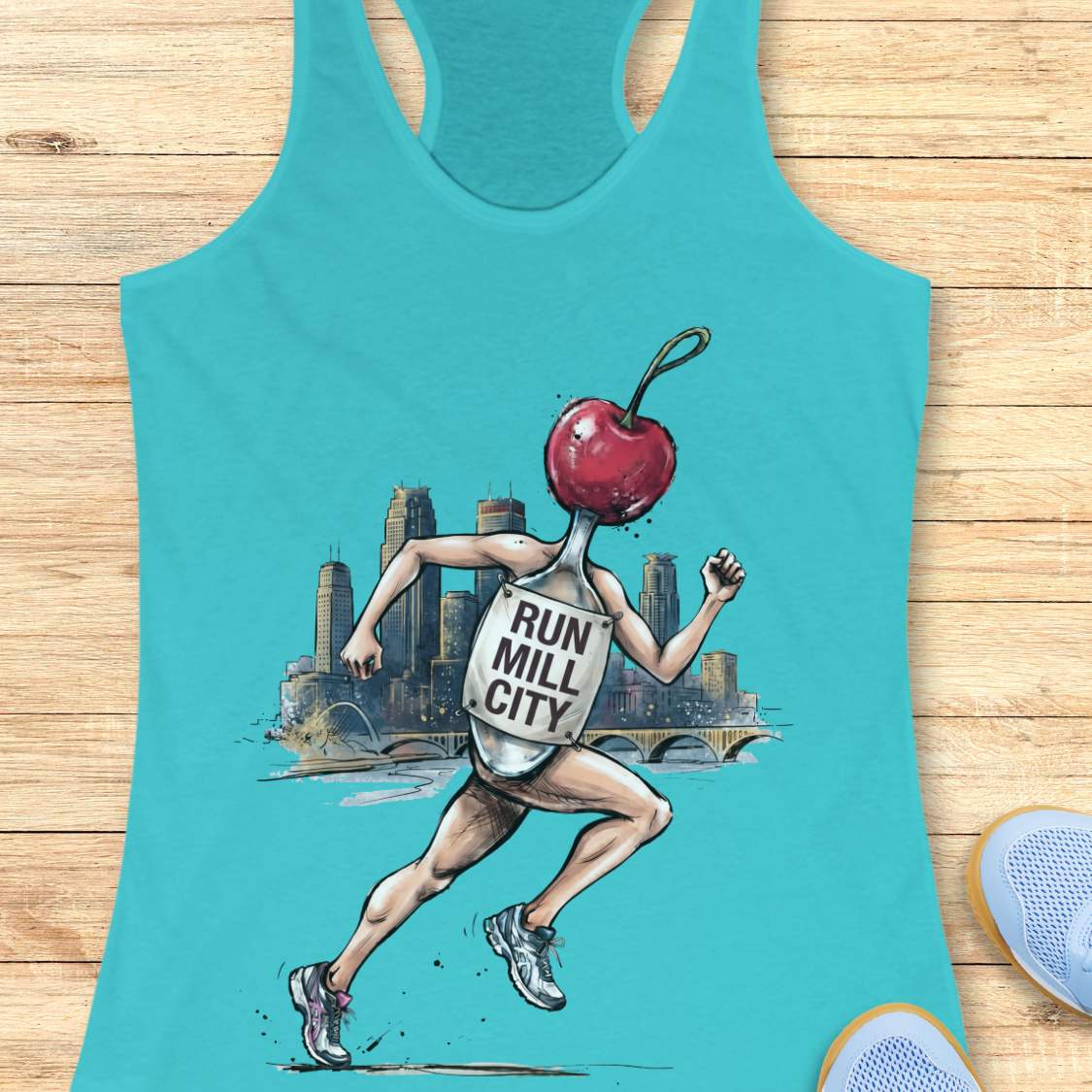 A turquoise racerback tank top laid flat on a light wood surface, featuring a graphic of a runner whose head is a red cherry and whose back displays the words 'RUN MILL CITY.' A partial sneaker appears at the bottom right corner.