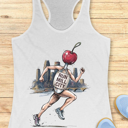 A white racerback tank top laid flat on a light wood plank background, featuring a graphic print of a runner with a red cherry for a head and the words 'RUN MILL CITY' on the back. Partial blue shoes are visible at the bottom right corner.