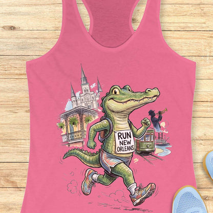 A pink racerback tank top laid flat on a light wood surface, featuring a colorful cartoon graphic of an alligator wearing a 'RUN NEW ORLEANS' shirt, with New Orleans landmarks and a streetcar in the background.