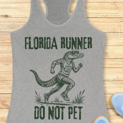 A grey racerback tank top laid flat on a light wood background, featuring a dark green graphic of an alligator running with the text 'FLORIDA RUNNER' above and 'DO NOT PET' below.