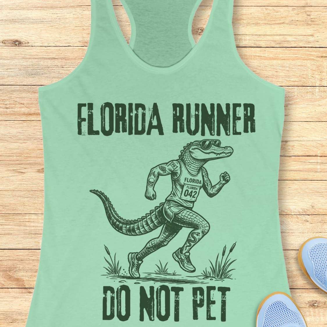 A mint green racerback tank top laid flat on a light wood background, featuring a dark green graphic of an alligator running with the text 'FLORIDA RUNNER' above and 'DO NOT PET' below.