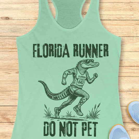 A mint green racerback tank top laid flat on a light wood background, featuring a dark green graphic of an alligator running with the text 'FLORIDA RUNNER' above and 'DO NOT PET' below.