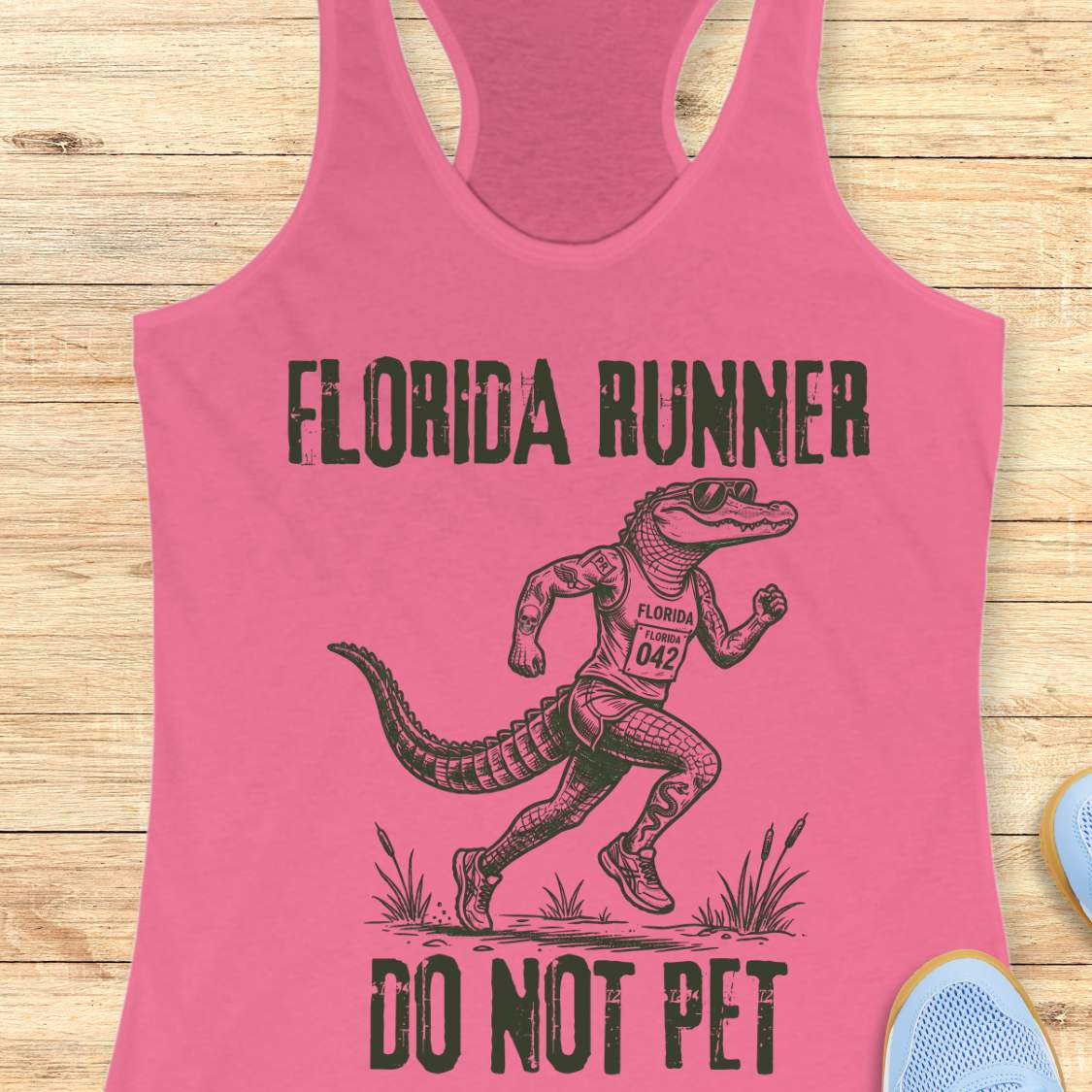 A pink racerback tank top laid flat on a light wood background, featuring a dark green graphic of an alligator running with the text 'FLORIDA RUNNER' above and 'DO NOT PET' below.