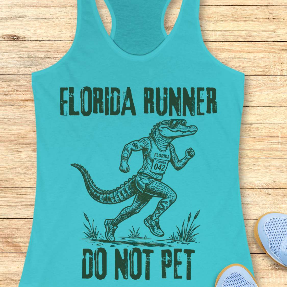 A blue  racerback tank top laid flat on a light wood background, featuring a dark green graphic of an alligator running with the text 'FLORIDA RUNNER' above and 'DO NOT PET' below.