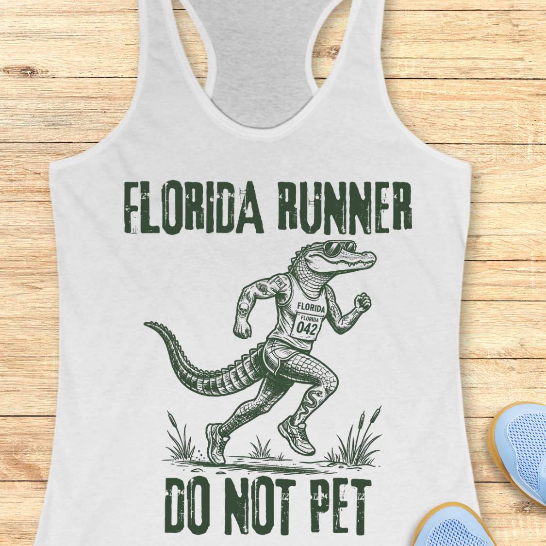 A white racerback tank top laid flat on a light wood background, featuring a dark green graphic of an alligator running with the text 'FLORIDA RUNNER' above and 'DO NOT PET' below.