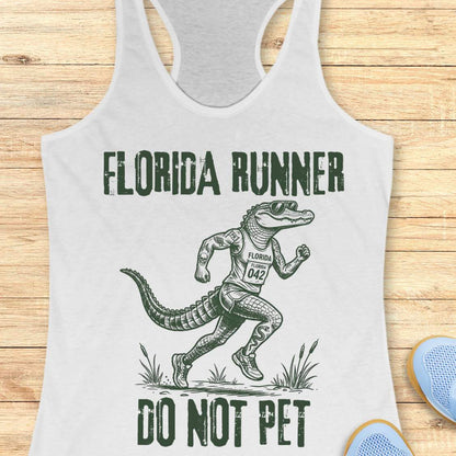 A white racerback tank top laid flat on a light wood background, featuring a dark green graphic of an alligator running with the text 'FLORIDA RUNNER' above and 'DO NOT PET' below.