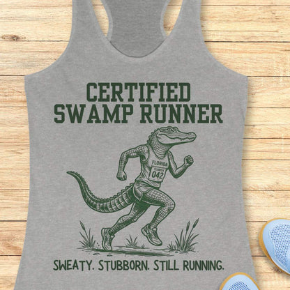 Funny swamp runner grey tank top featuring a running alligator wearing a race bib and athletic gear with the slogan “Certified Swamp Runner: Sweaty. Stubborn. Still Running.” Perfect for women runners, Florida runners, trail runners, marathon training, and runners who embrace hot, humid miles.
