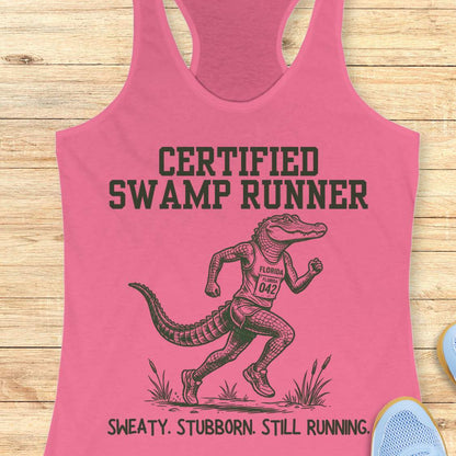 Funny swamp runner pink tank top featuring a running alligator wearing a race bib and athletic gear with the slogan “Certified Swamp Runner: Sweaty. Stubborn. Still Running.” Perfect for women runners, Florida runners, trail runners, marathon training, and runners who embrace hot, humid miles.