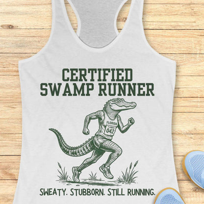 Funny swamp runner white tank top featuring a running alligator wearing a race bib and athletic gear with the slogan “Certified Swamp Runner: Sweaty. Stubborn. Still Running.” Perfect for women runners, Florida runners, trail runners, marathon training, and runners who embrace hot, humid miles.