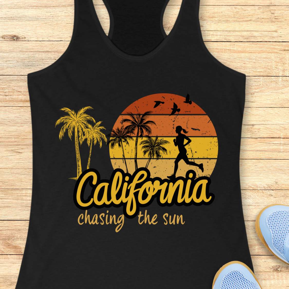A black racerback tank top laid flat on a light wood background, featuring a distressed sunset graphic with palm trees, a running silhouette, and the text 'California chasing the sun.' Partial blue sneakers are visible at the bottom right as styling props.