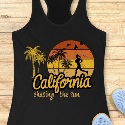 A black racerback tank top laid flat on a light wood background, featuring a distressed sunset graphic with palm trees, a running silhouette, and the text 'California chasing the sun.' Partial blue sneakers are visible at the bottom right as styling props.