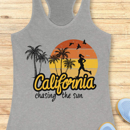 A heather gray racerback tank top laid flat on a light wood background, featuring a colorful graphic print with palm trees, a running silhouette, birds, a sunset gradient, and the words 'California chasing the sun.'.