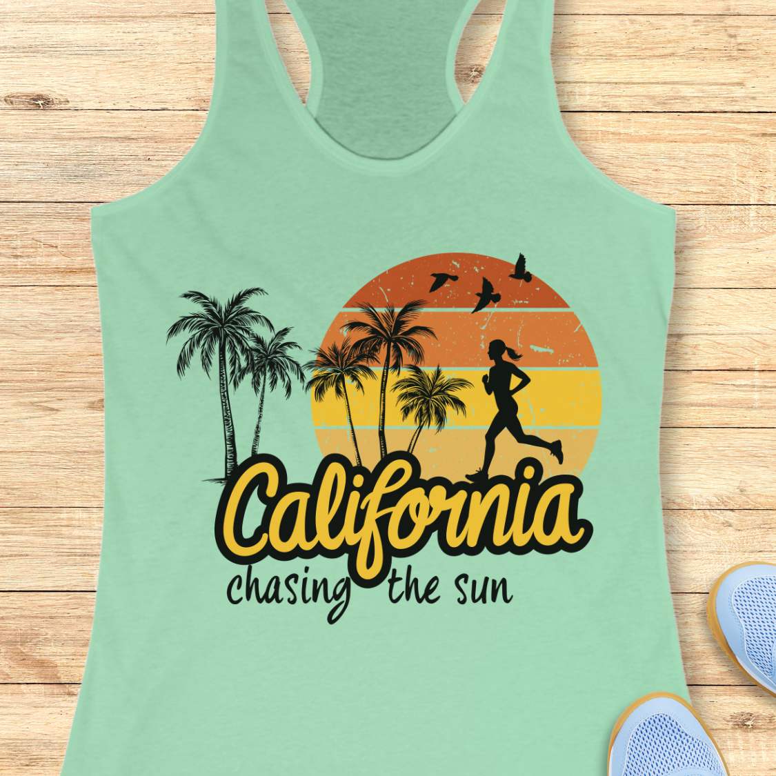 A mint green racerback tank top laid flat on a light wood background, featuring a retro graphic with palm trees, a running silhouette, a sunset circle, and the text 'California chasing the sun.' Partial blue sneakers are visible at the bottom right as styling props.