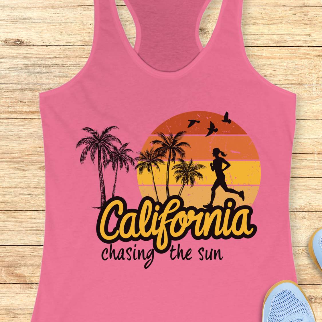A pink racerback tank top laid flat on a light wood background, featuring a retro sunset graphic with palm trees, a running silhouette, and the text 'California chasing the sun.' Partial blue shoes are visible at the edges.