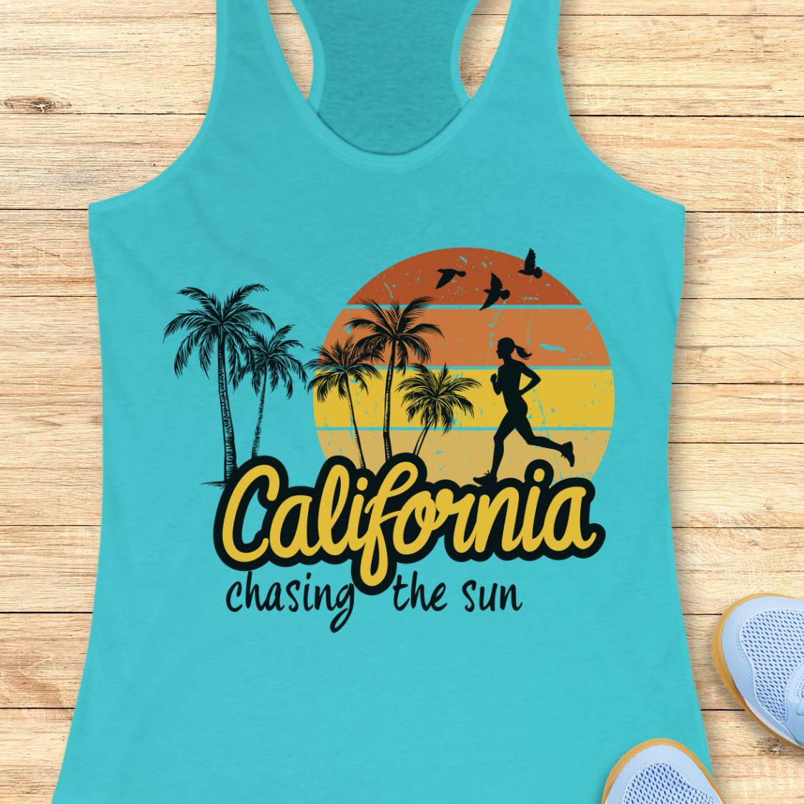 A turquoise racerback tank top laid flat on a light wood surface, featuring a retro sunset graphic with palm trees, a running silhouette, birds, and the text 'California chasing the sun.' Partial sneakers are visible at the edges as styling props.