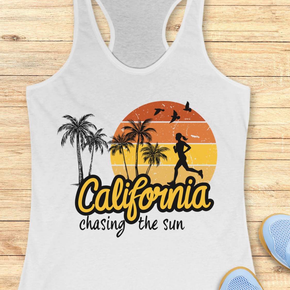 A white racerback tank top laid flat on a light wood background, featuring a retro-style graphic with palm trees, a running silhouette, a sunset circle, and the text 'California chasing the sun.' Partial blue shoes are visible at the bottom right as styling props.