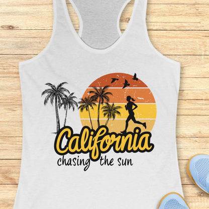 A white racerback tank top laid flat on a light wood background, featuring a retro-style graphic with palm trees, a running silhouette, a sunset circle, and the text 'California chasing the sun.' Partial blue shoes are visible at the bottom right as styling props.