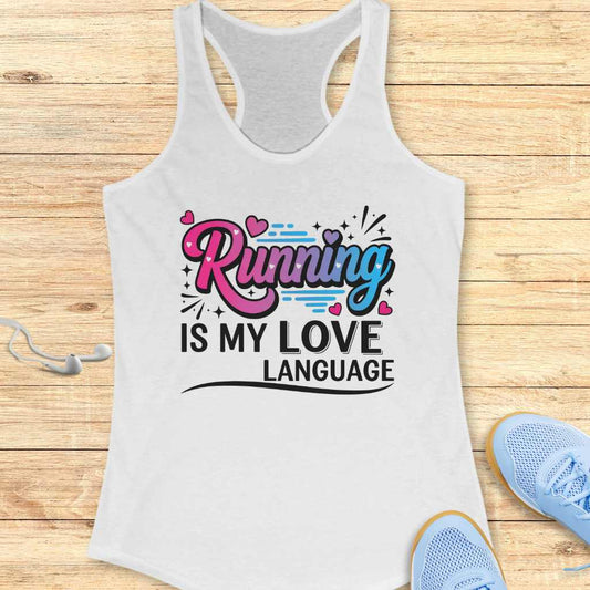 White women's racerback tank top with "Running IS MY LOVE LANGUAGE" slogan in vibrant pink and blue gradient lettering with heart and dash accents, displayed on a light wooden surface beside white wired earbuds and the toes of light blue running shoes