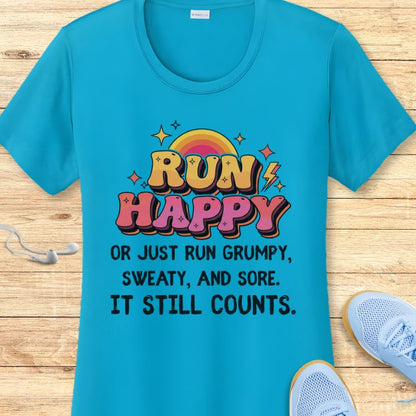 Turquoise blue running t-shirt on a wooden surface, prominently displaying the vibrant 'RUN HAPPY' text with a rainbow and lightning bolt, along with the motivational slogan 'OR JUST RUN GRUMPY, SWEATY, AND SORE IT STILL COUNTS', framed by white earbuds and light blue running shoes