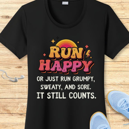 Photograph of a black t-shirt displaying a vibrant retro-style RUN HAPPY graphic in orange-yellow and pink-red gradient letters with lightning bolts and stars above a rainbow sun, along with the complete motivational phrase OR JUST RUN GRUMPY SWEATY AND SORE IT STILL COUNTS, all resting on a rustic wooden background next to white earbuds and bright blue running shoes