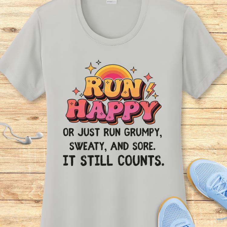 Product photo of a light grey t-shirt featuring a vibrant retro-style 'RUN HAPPY' graphic in orange and pink with a rainbow sun and lightning bolts, above the black text 'OR JUST RUN GRUMPY, SWEATY, AND SORE. IT STILL COUNTS', with white earbuds and light blue running shoes visible on a wooden floor
