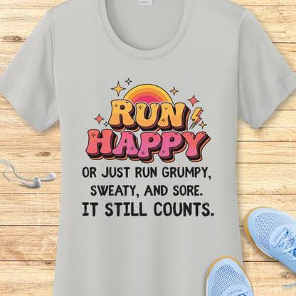 Product photo of a light grey t-shirt featuring a vibrant retro-style 'RUN HAPPY' graphic in orange and pink with a rainbow sun and lightning bolts, above the black text 'OR JUST RUN GRUMPY, SWEATY, AND SORE. IT STILL COUNTS', with white earbuds and light blue running shoes visible on a wooden floor