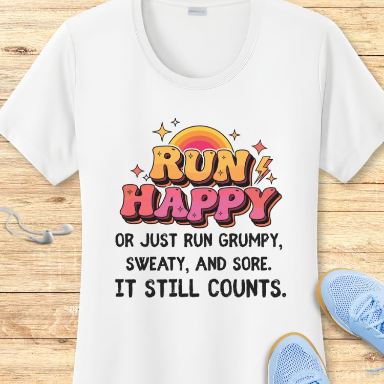 White T-shirt with a retro orange and pink RUN HAPPY slogan featuring a rainbow and lightning bolts, alongside black text stating running grumpy sweaty or sore still counts, positioned with white earbuds and light blue running shoes on a wooden background
