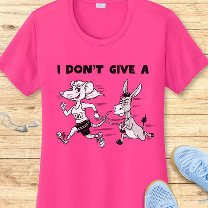 Graphic print on a hot pink t-shirt featuring a cartoon mouse with a 101 bib running and pulling a grumpy donkey on a leash, with the text I DON'T GIVE A above, displayed on a wooden surface with blue sneakers and white earbuds nearby