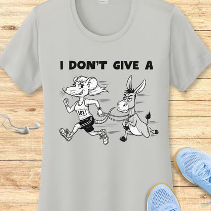 Light gray t-shirt featuring a cartoon of a smiling mouse runner with bib 101 pulling a grumpy donkey on a leash, beneath the text I DON'T GIVE A, shown on a wooden background with light blue sneakers