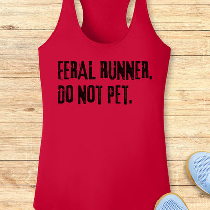A red racerback performance tank top with black distressed text reading 'FERAL RUNNER, DO NOT PET.' laid flat on a light wood plank background, with partial blue sneakers visible at the bottom right.