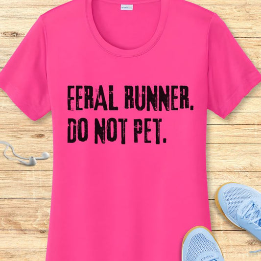 Product photo of a vibrant fuchsia pink t-shirt featuring distressed black text FERAL RUNNER DO NOT PET displayed on a light wooden surface with light blue running shoes and white wired earbuds partially visible