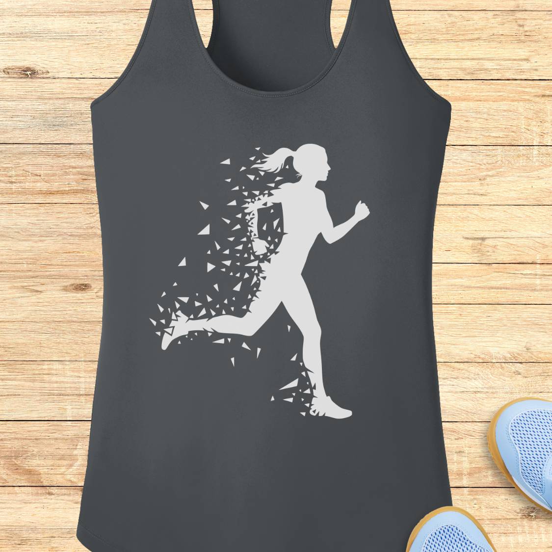 A dark charcoal gray racerback tank top featuring a white graphic of a running woman whose body dissolves into small geometric shapes. The tank top is displayed flat on a light-colored wooden plank background, with the tips of blue sneakers partially visible in the bottom right corner.