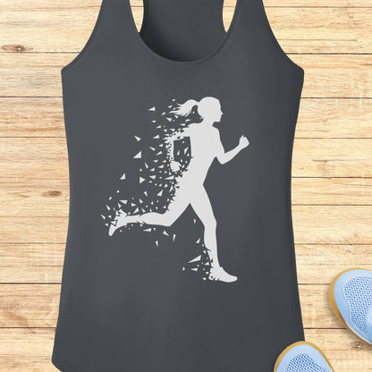 A dark charcoal gray racerback tank top featuring a white graphic of a running woman whose body dissolves into small geometric shapes. The tank top is displayed flat on a light-colored wooden plank background, with the tips of blue sneakers partially visible in the bottom right corner.
