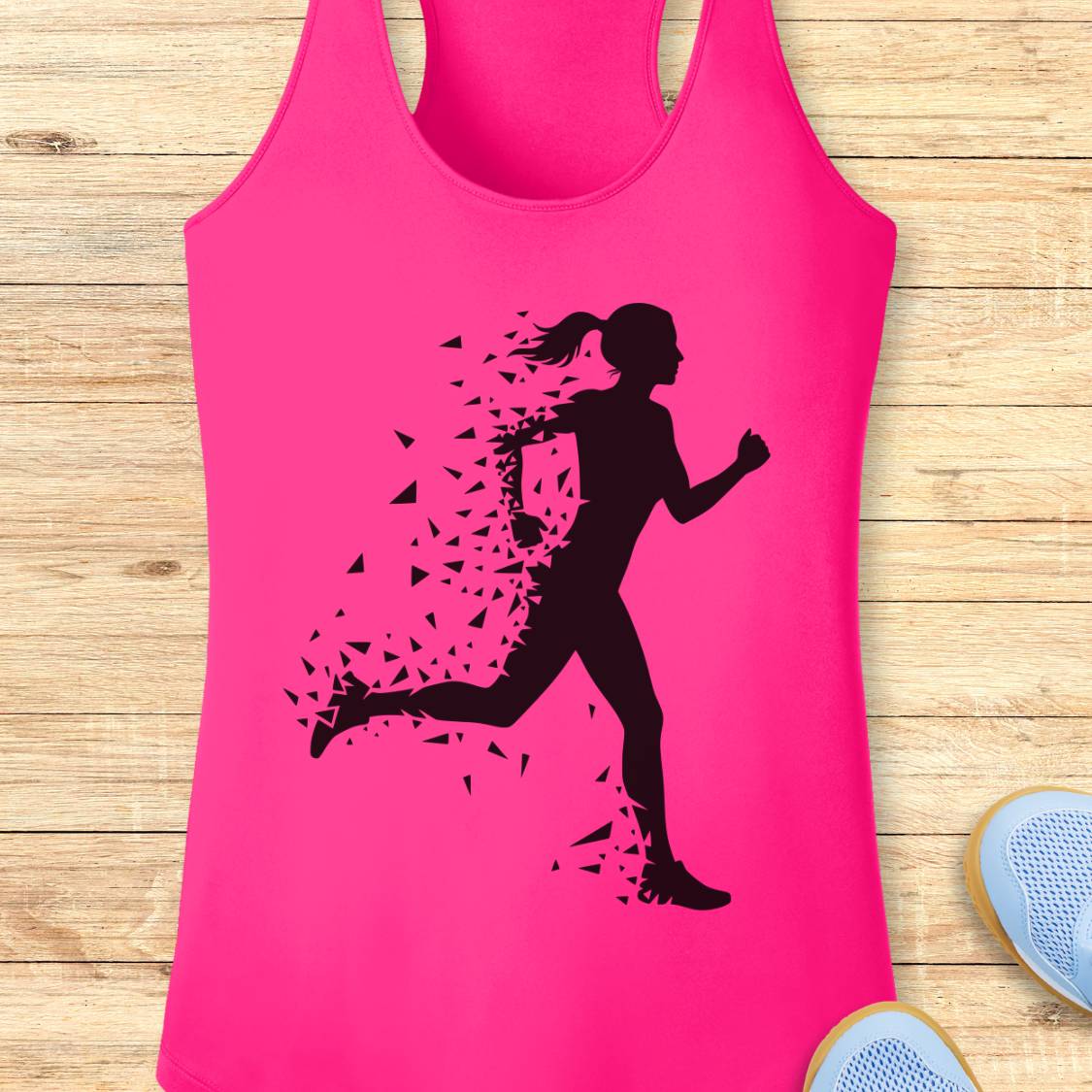 A bright pink racerback tank top with a black silhouette of a running woman who appears to be breaking apart into small fragments, laid flat on a light wood plank background with partial shoes visible at the edges.