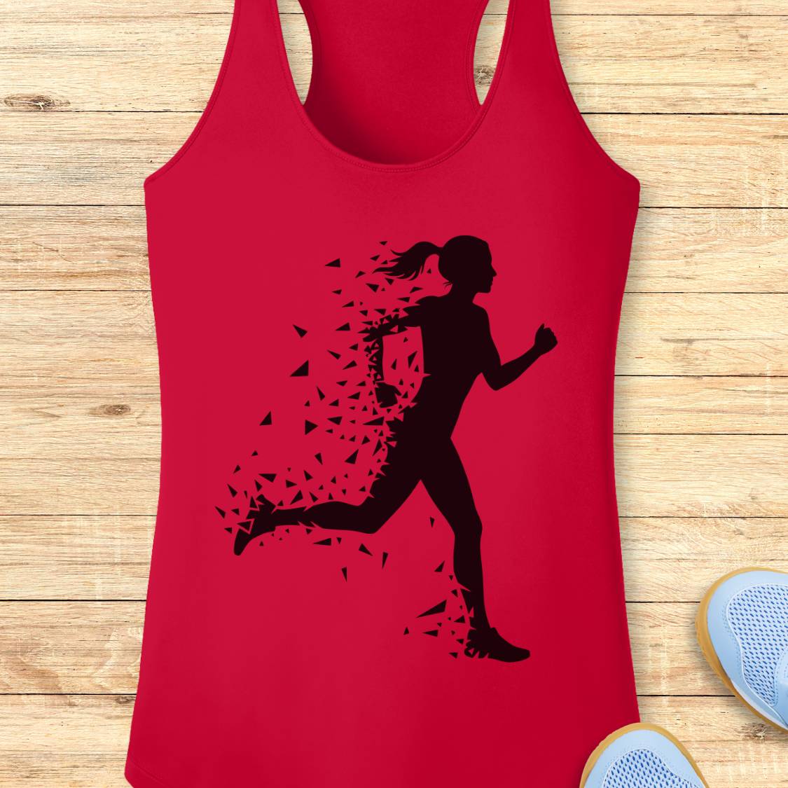 A red racerback tank top with a black silhouette graphic of a running woman whose body dissolves into small fragments, displayed flat on a light wood plank background with partial sneakers visible at the edges.