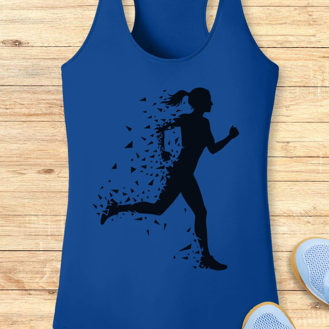 A royal blue racerback tank top featuring a black silhouette graphic of a running woman dissolving into small fragments, laid flat on a light wood plank background with partial sneakers visible at the bottom right.