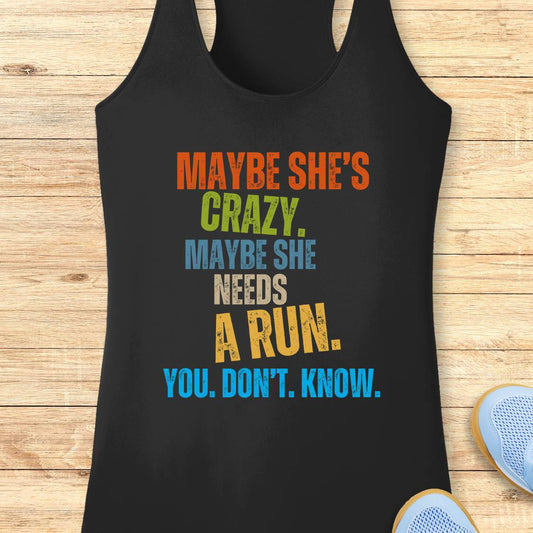 A black racerback performance tank top laid flat on a light wood surface, featuring a colorful distressed text graphic that reads, 'MAYBE SHE'S CRAZY. MAYBE SHE NEEDS A RUN.