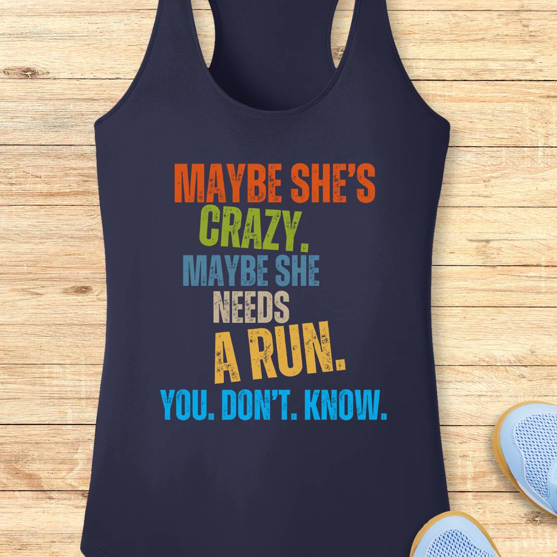 A navy blue performance racerback tank top with a colorful motivational text graphic reading 'MAYBE SHE'S CRAZY. MAYBE SHE NEEDS A RUN.