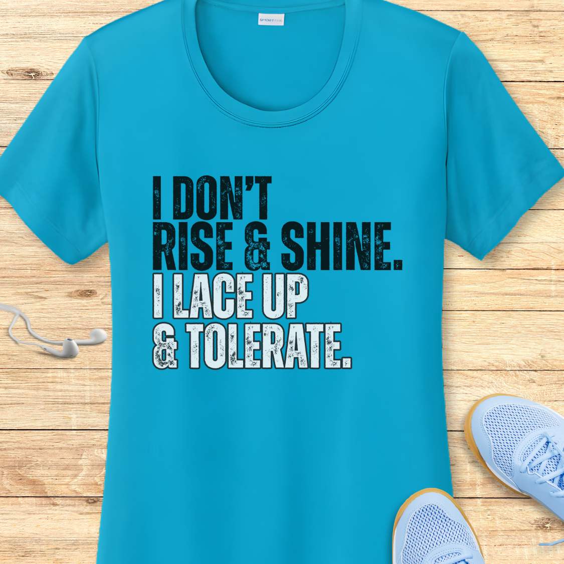 Blue t-shirt with humorous text 'I DON'T RISE & SHINE. I LACE UP & TOLERATE.'  on a wooden surface with sneakers.