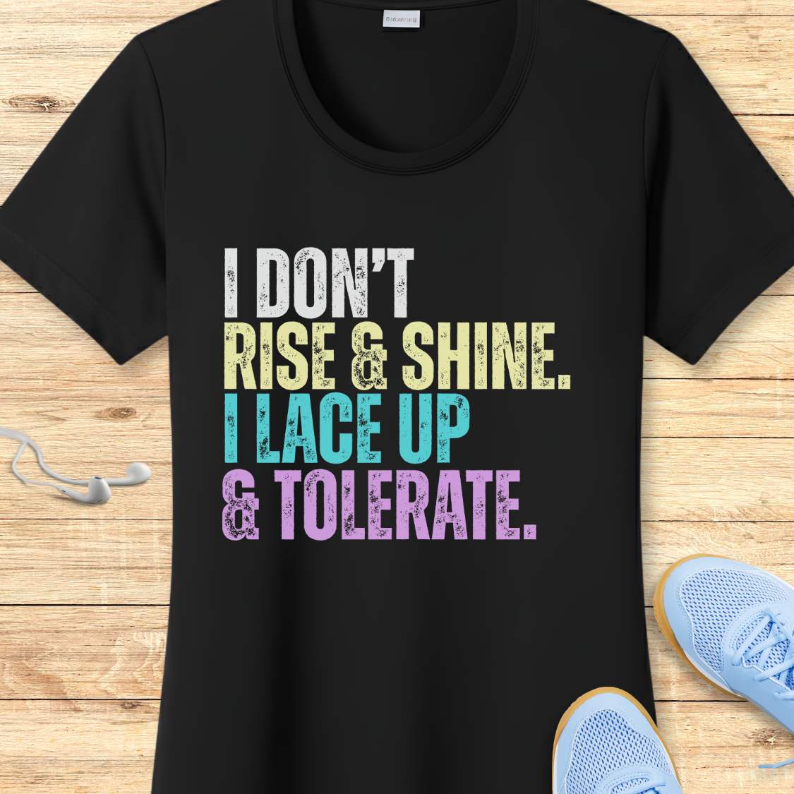 Black t-shirt with colorful text 'I DON'T RISE & SHINE. I LACE UP & TOLERATE.'  on a wooden surface with sneakers.