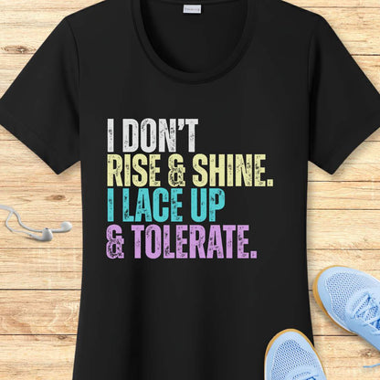 Black t-shirt with colorful text 'I DON'T RISE & SHINE. I LACE UP & TOLERATE.'  on a wooden surface with sneakers.