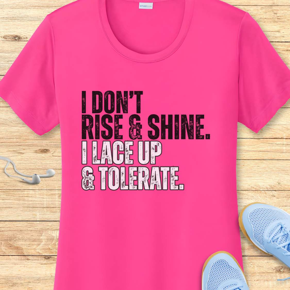 Pink t-shirt with text 'I DON'T RISE & SHINE. I LACE UP & TOLERATE.' on a wooden surface with sneakers
