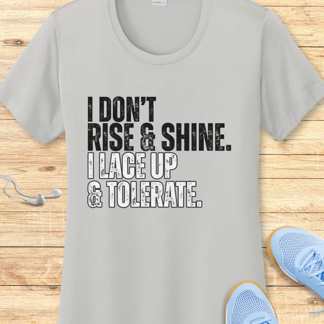 Gray t-shirt with text 'I DON'T RISE & SHINE. I LACE UP & TOLERATE.' on a wooden surface with blue sneakers.