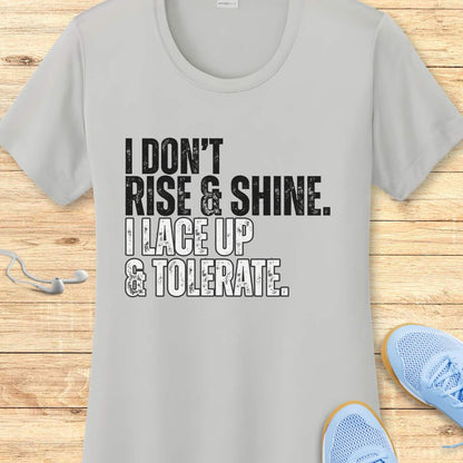 Gray t-shirt with text 'I DON'T RISE & SHINE. I LACE UP & TOLERATE.' on a wooden surface with blue sneakers.