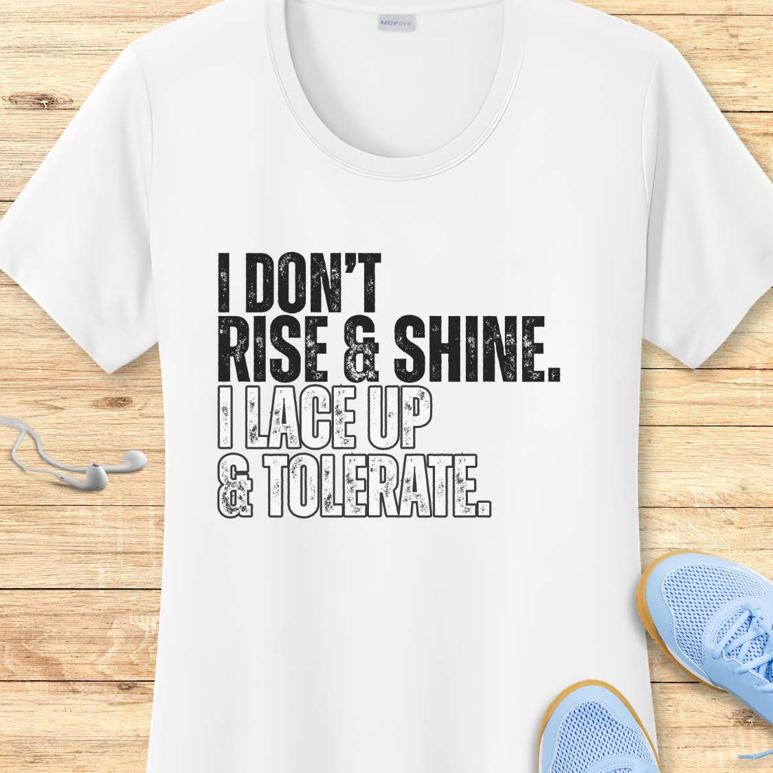 White t-shirt with humorous text 'I DON'T RISE & SHINE. I LACE UP & TOLERATE.'  on a wooden surface with sneakers.
