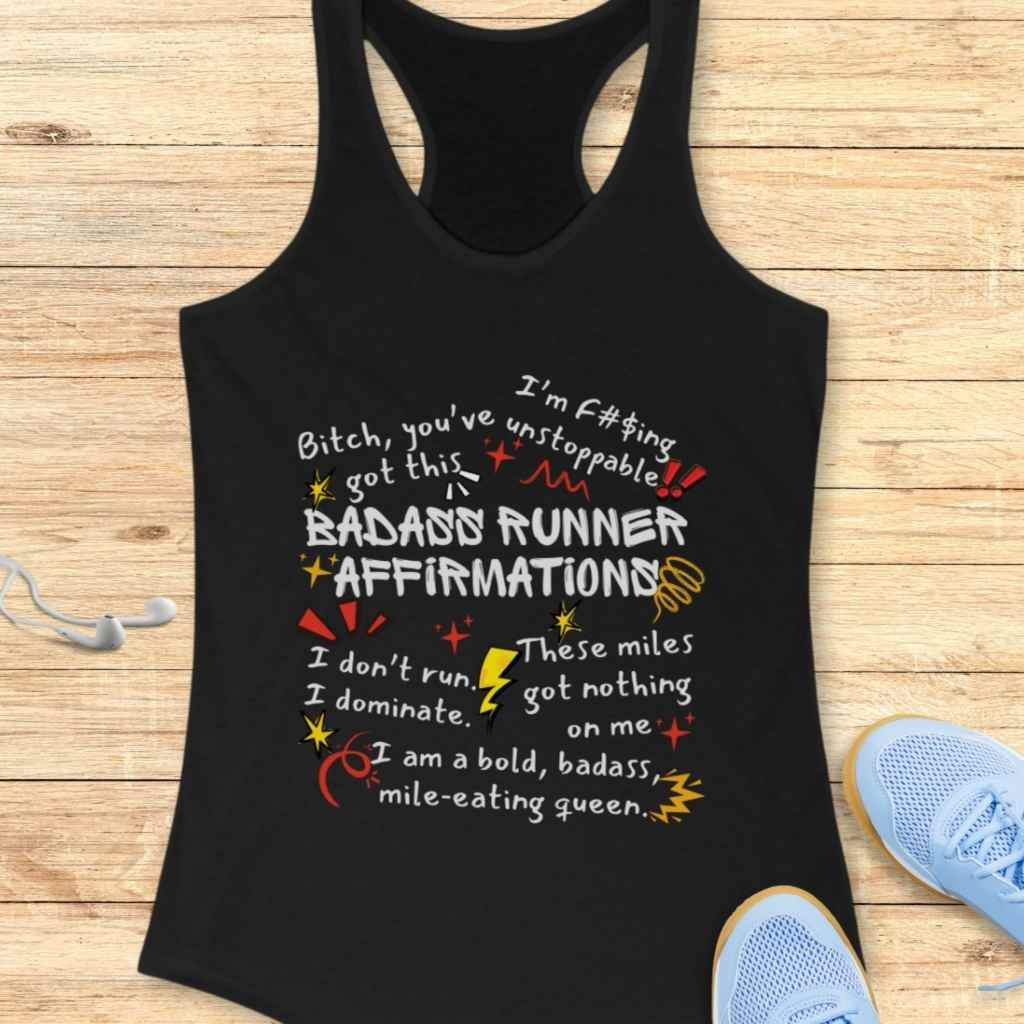 Black cotton tank top printed with "Badass Runner Affirmations" and empowering phrases like "Bitch, you've got this" and "I'm f#$ing unstoppable" in white, yellow, and red text with stars and lightning bolt graphics, displayed on a wooden surface next to light blue running shoes and white earbuds