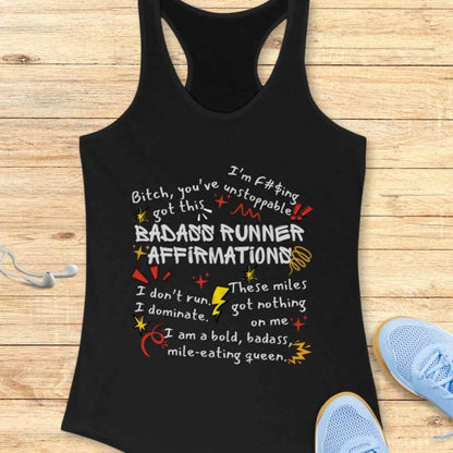 Black cotton tank top printed with "Badass Runner Affirmations" and empowering phrases like "Bitch, you've got this" and "I'm f#$ing unstoppable" in white, yellow, and red text with stars and lightning bolt graphics, displayed on a wooden surface next to light blue running shoes and white earbuds