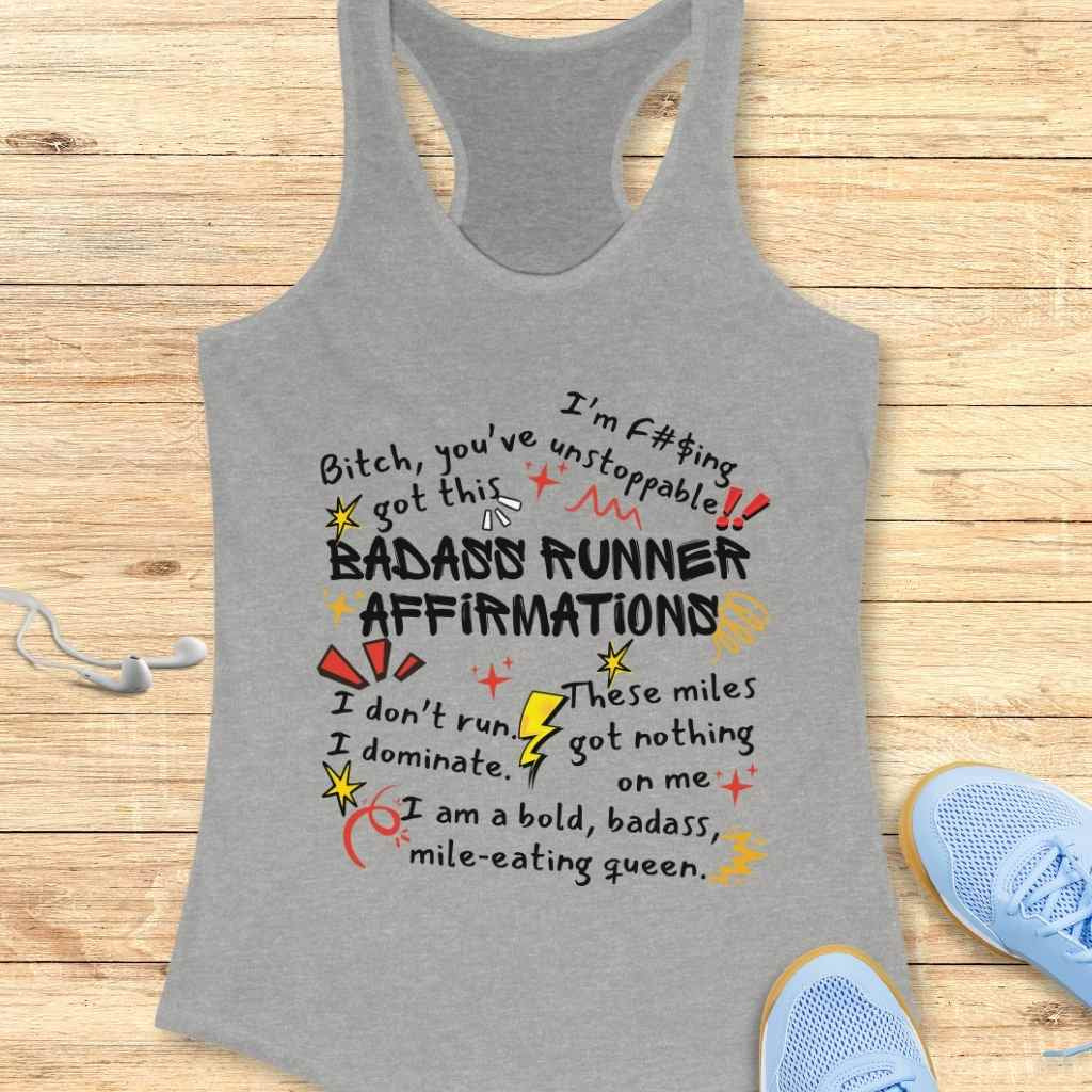 Graphic design on a grey racerback tank top featuring colorful hand-drawn typography with bold 'BADASS RUNNER AFFIRMATIONS' and empowering phrases like 'Bitch, you've got this' and 'I am a bold, badass, mile-eating queen' displayed on a wooden surface with white earbuds and light blue running shoes