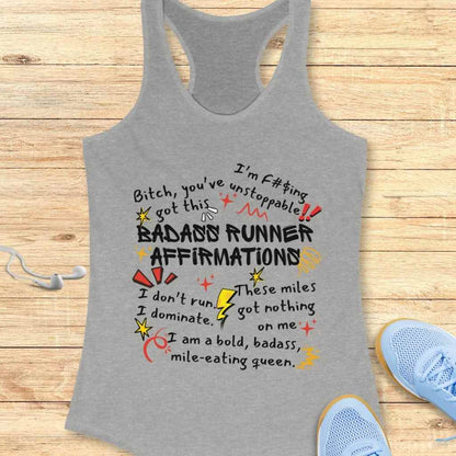Graphic design on a grey racerback tank top featuring colorful hand-drawn typography with bold 'BADASS RUNNER AFFIRMATIONS' and empowering phrases like 'Bitch, you've got this' and 'I am a bold, badass, mile-eating queen' displayed on a wooden surface with white earbuds and light blue running shoes